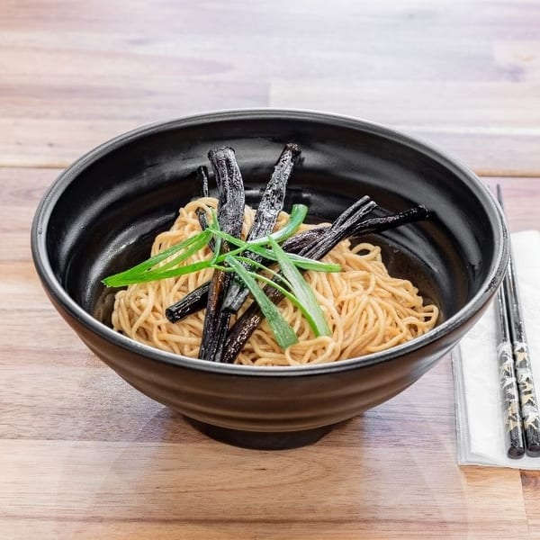 Black bowl of noodles with dark strips and green onions, chopsticks on a napkin beside it.