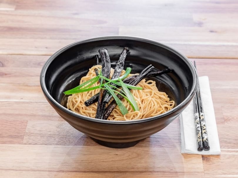 Black bowl of noodles with dark strips and green onions, chopsticks on a napkin beside it.