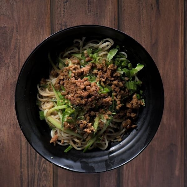 Overhead view of a black bowl filled with noodles, ground meat, and green vegetables on a wooden table.