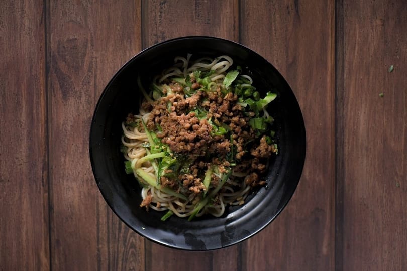 Overhead view of a black bowl filled with noodles, ground meat, and green vegetables on a wooden table.