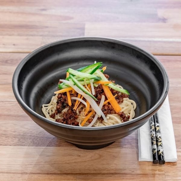 Black bowl of noodles with minced meat, shredded carrots, cucumbers, and bean sprouts on a wooden table.