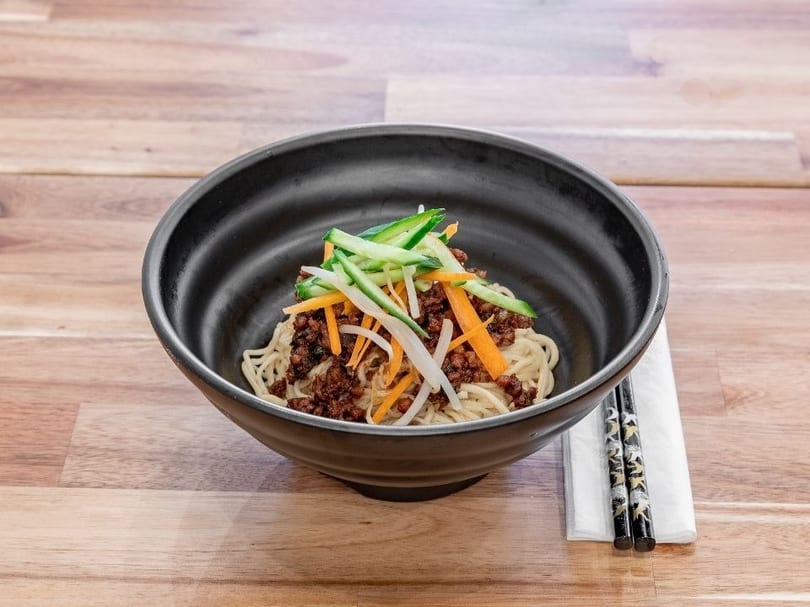 Black bowl of noodles with minced meat, shredded carrots, cucumbers, and bean sprouts on a wooden table.