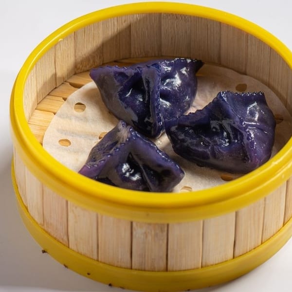 Three vibrant purple dumplings rest on parchment paper inside a bamboo steamer with a yellow rim.