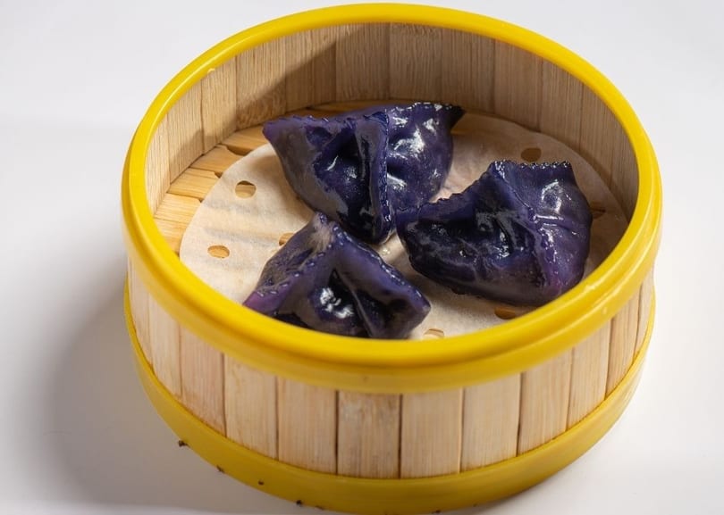 Three vibrant purple dumplings rest on parchment paper inside a bamboo steamer with a yellow rim.