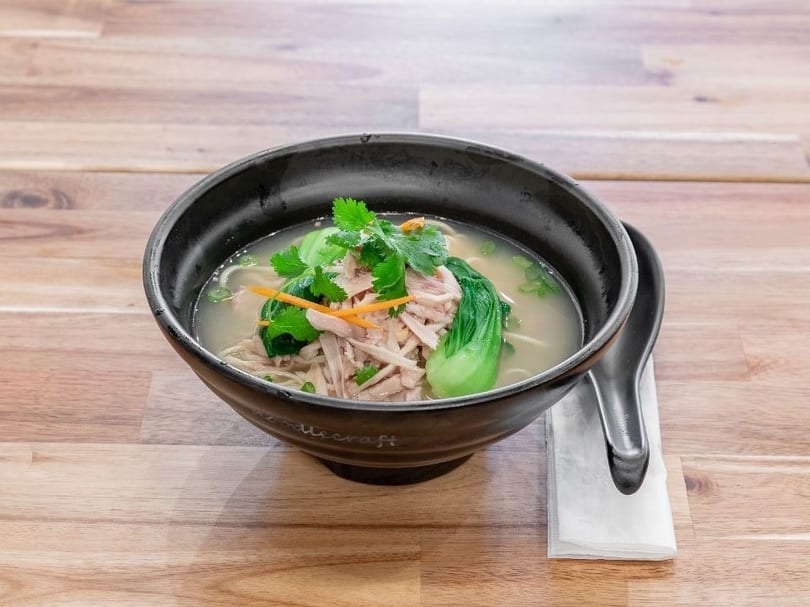 Chicken noodle soup with bok choy, cilantro, and shredded carrots in a black bowl.