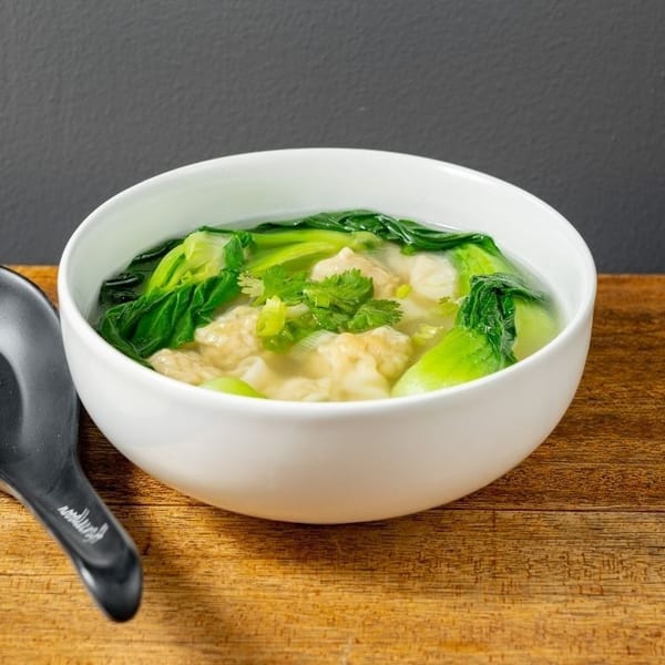 Wonton soup with bok choy and cilantro in a white bowl with a black spoon on a wooden table.