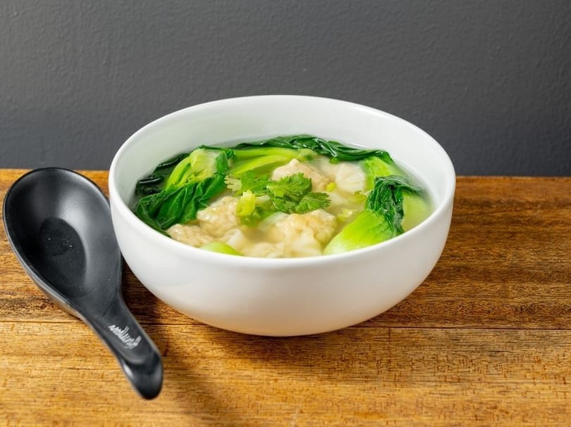 Wonton soup with bok choy and cilantro in a white bowl with a black spoon on a wooden table.