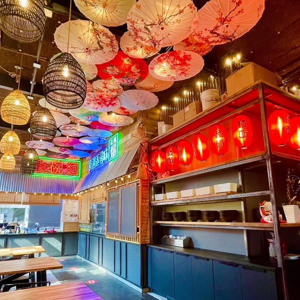 Restaurant interior with hanging paper umbrellas, woven light fixtures, red lanterns, and a neon sign.