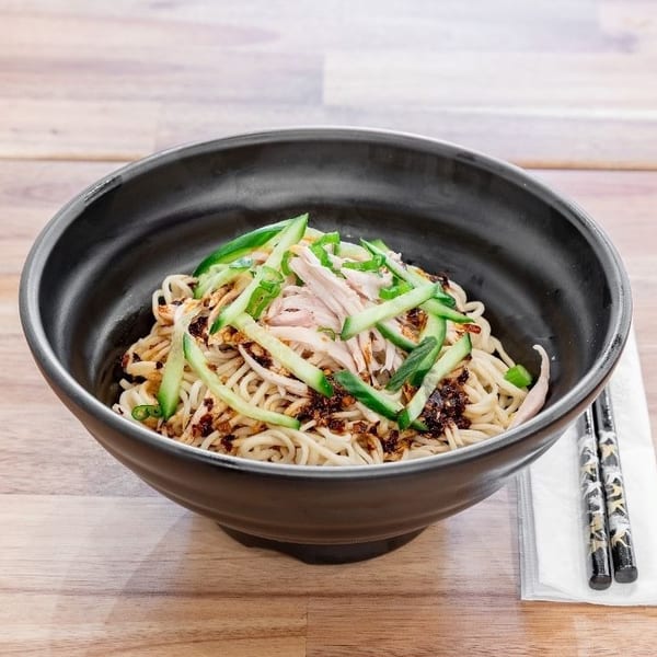 A black bowl of noodles with shredded chicken, cucumber, and chili oil on a wooden table.
