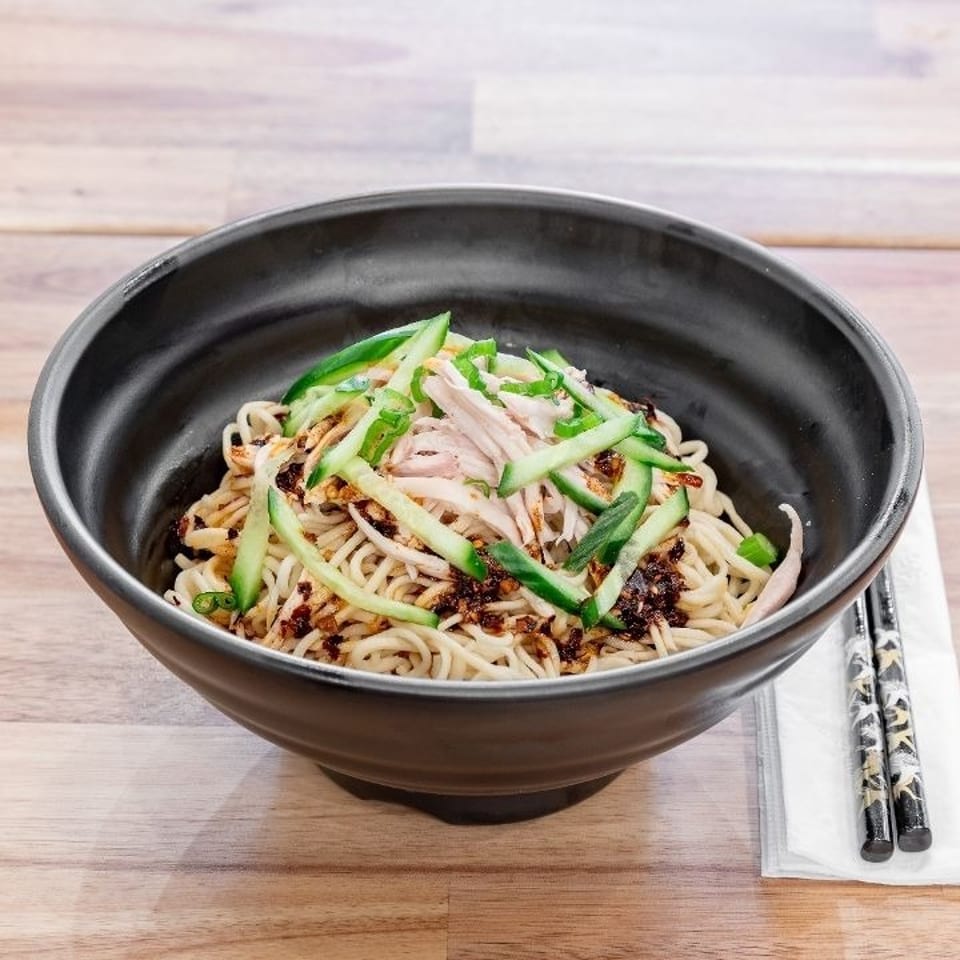 A black bowl of noodles with shredded chicken, cucumber, and chili oil on a wooden table.