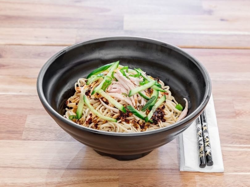 A black bowl of noodles with shredded chicken, cucumber, and chili oil on a wooden table.