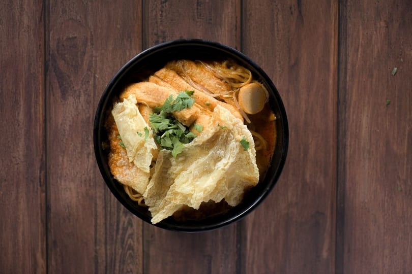 Overhead view of a black bowl filled with curry noodles, tofu, fish cakes, and cilantro.