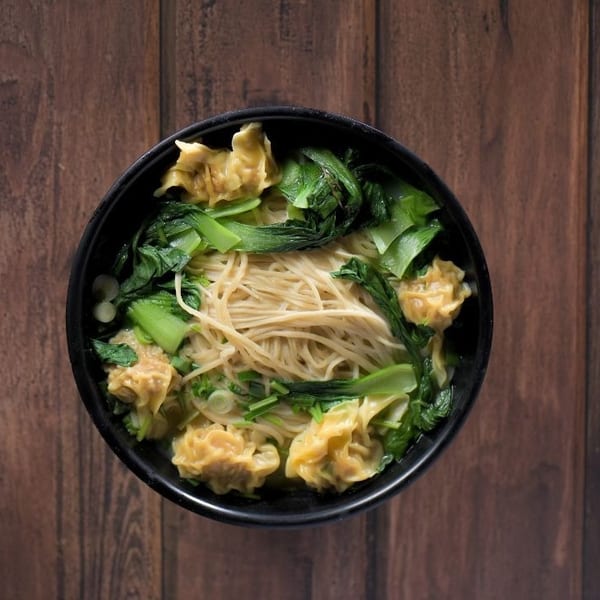 Overhead view of a black bowl filled with noodles, wontons, and green vegetables on a wooden table.