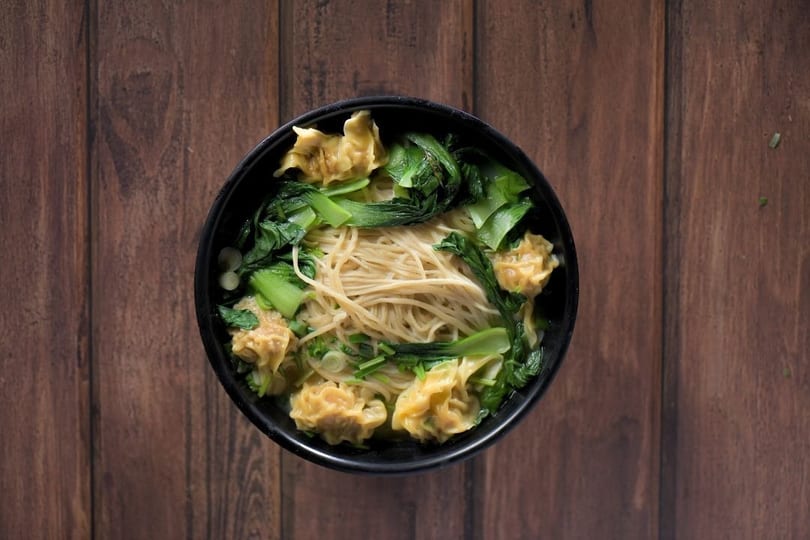 Overhead view of a black bowl filled with noodles, wontons, and green vegetables on a wooden table.
