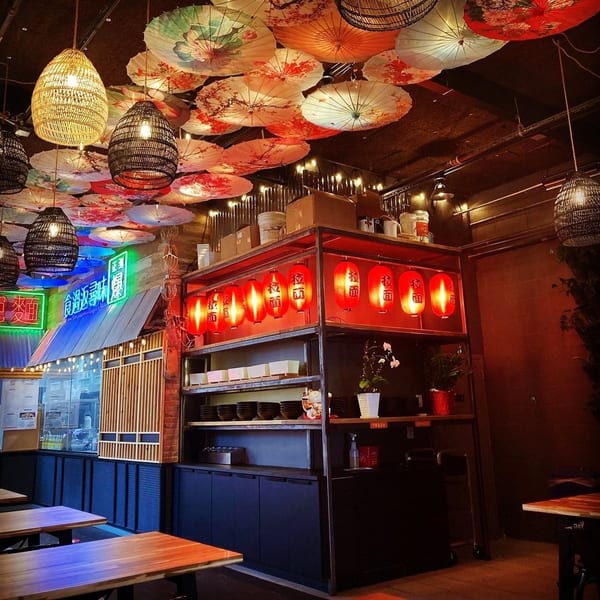 Restaurant interior with hanging paper umbrellas, woven lamps, and red lanterns over shelves.