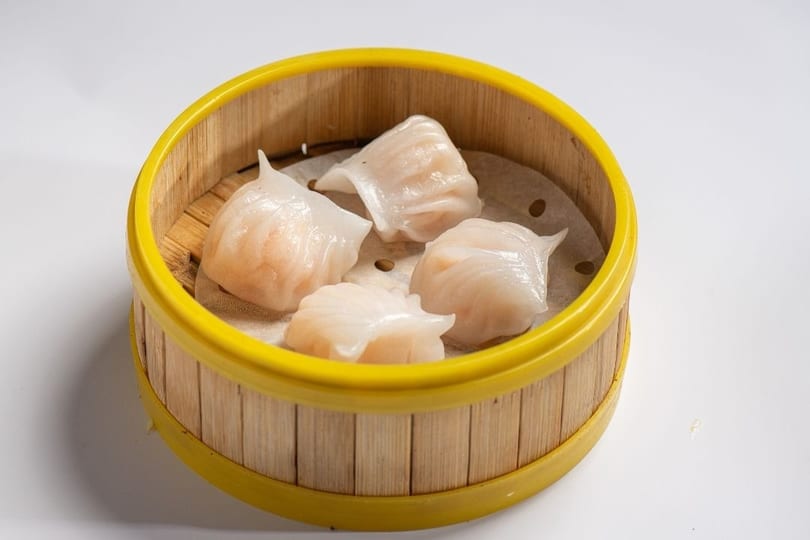 Four translucent shrimp dumplings (har gow) in a bamboo steamer basket on a white background.
