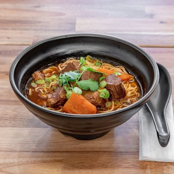 A black bowl of beef noodle soup with carrots, green onions, and cilantro on a wooden table.