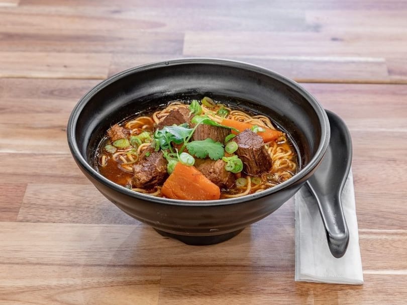 A black bowl of beef noodle soup with carrots, green onions, and cilantro on a wooden table.