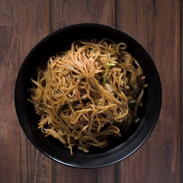 Overhead view of a black bowl filled with stir-fried noodles and vegetables on a wooden table.
