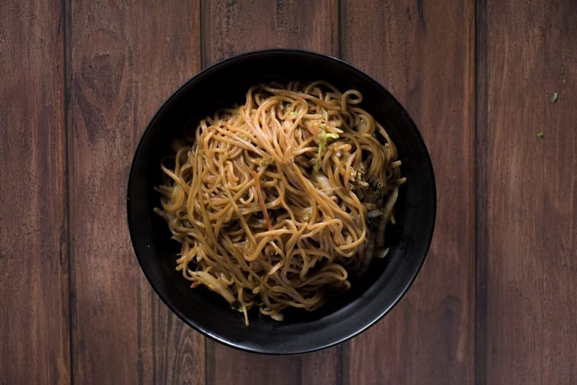 Overhead view of a black bowl filled with stir-fried noodles and vegetables on a wooden table.