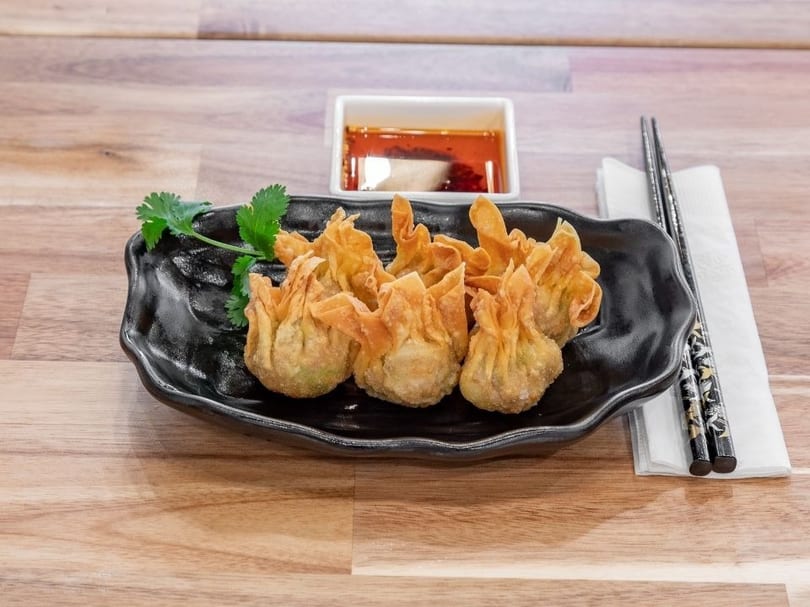 Crispy fried wontons on a black plate with cilantro, chili oil, and chopsticks on a wooden table.